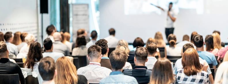 Large audience seated facing a presenter at a whiteboard during a lecture or conference presentation