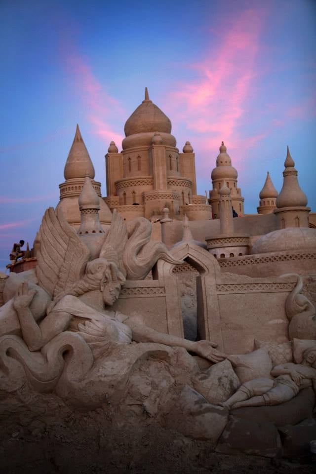 Elaborate sand castle with towering domes and spires against a twilight sky with pink clouds and detailed dragon sculpture in foreground