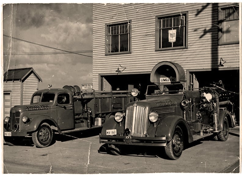 Vintage fire trucks parked in front of a brick firehouse building, black and white historical photograph