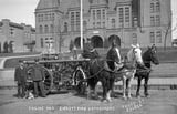 Horse-drawn fire engine with three horses and firefighters in front of a large brick building, early 1900s
